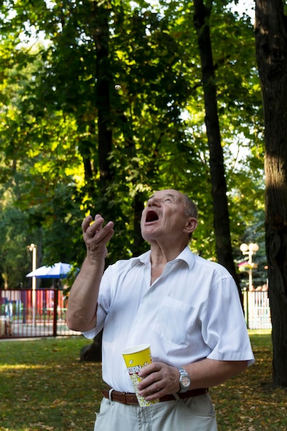 Un uomo anziano cammina da solo nel parco in estate Il pensionato lancia allegramente popcorn da un bicchiere e lo prende con la bocca spalancata