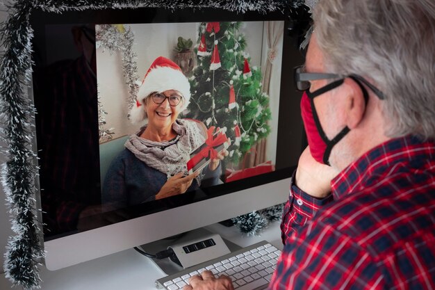 Un uomo anziano che indossa una maschera a causa del coronavirus in videochiamata per gli auguri di Natale da casa con sua moglie lontana