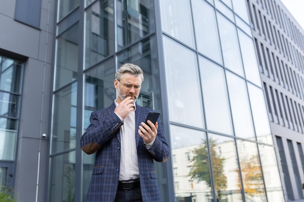 Un uomo anziano dai capelli grigi in giacca e occhiali si trova fuori da un ufficio un grattacielo guarda preoccupato