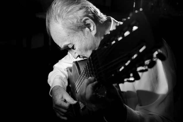 Un uomo anziano in camicia bianca che suona una chitarra acustica. Sfondo scuro. Monocromo.