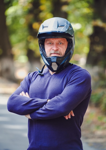 Un uomo anziano in casco scuro all'aperto. Vestiti casuali. Mani incrociate. Maglione blu. Sfondo sfocato all'aperto. Avvicinamento.