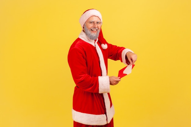 Un uomo anziano sorridente felice con la barba grigia che indossa il costume di babbo natale apre una busta con una lettera, riceve una busta di congratulazioni con il Natale. Colpo dello studio dell'interno isolato su priorità bassa gialla.