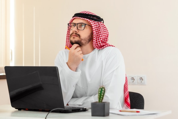 Un uomo arabo, un uomo d'affari, uno sceicco in costume arabo nazionale lavora a un tavolo in ufficio. Investimenti, affari, lavoro via Internet, contratti online.