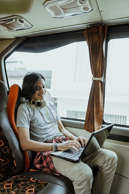 Un uomo asiatico con le cuffie e che usa un laptop durante la digitazione siede vicino a una finestra sull'autobus