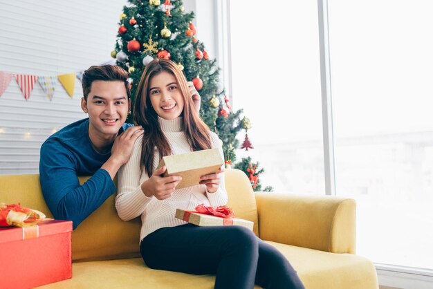 Un uomo asiatico felice sorprende la sua ragazza con regali di Natale a casa con un albero di Natale nell'immagine di sfondo con spazio per la copia