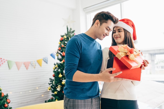 Un uomo asiatico felice sorprende la sua ragazza con regali di Natale a casa con un albero di Natale nell'immagine di sfondo con spazio per la copia