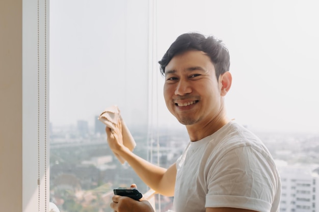 Un uomo asiatico thailandese felice pulisce il vetro della finestra in un appartamento con vista sulla città sorridendo guardando la telecamera