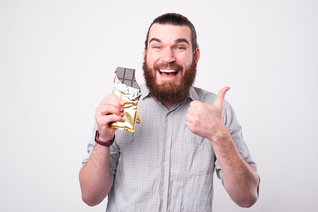 Un uomo barbuto allegro sta tenendo un cioccolato e sorridendo alla macchina fotografica sta mostrando un pollice in su e che ama il cioccolato