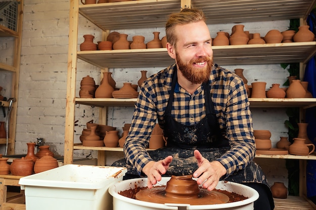 Un uomo barbuto che modella un vaso di argilla su un tornio da vasaio in un laboratorio di ceramica. vasaio, argilla, vaso, ceramica e vasaio. master e ceramiche.