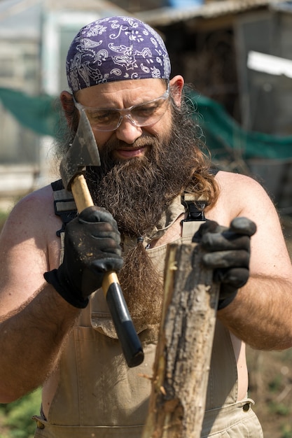Un uomo barbuto di mezza età con una bandana taglia tronchi con un'ascia Brutale in tuta fa il duro lavoro
