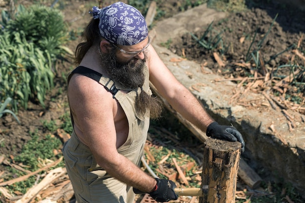 Un uomo barbuto di mezza età con una bandana taglia tronchi con un'ascia Brutale in tuta fa il duro lavoro