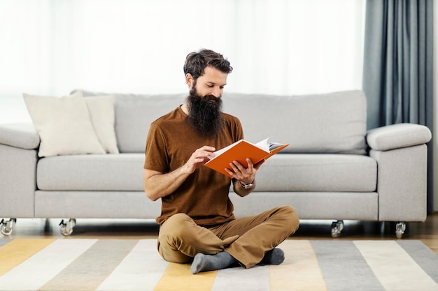 Un uomo barbuto seduto sul pavimento nella sua casa accogliente che si rilassa e legge un libro