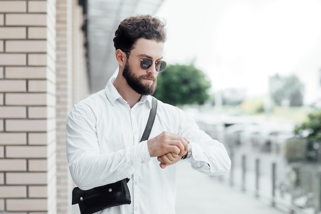 Un uomo barbuto, serio ed elegante guarda l'orologio e cammina per le strade della città vicino al moderno centro uffici