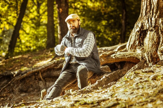 Un uomo barbuto serio seduto accanto a un albero nella foresta in una buona giornata