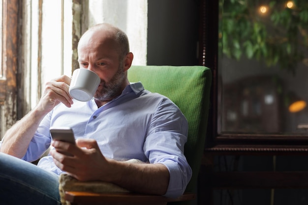 Un uomo beve un caffè e controlla le notizie al telefono seduto su una sedia vicino alla finestra
