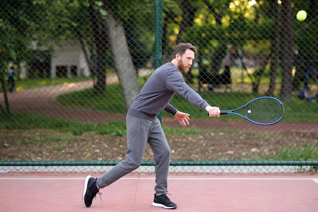 Un uomo caucasico barbuto con abbigliamento sportivo grigio che cerca di colpire la pallina da tennis sul campo da tennis