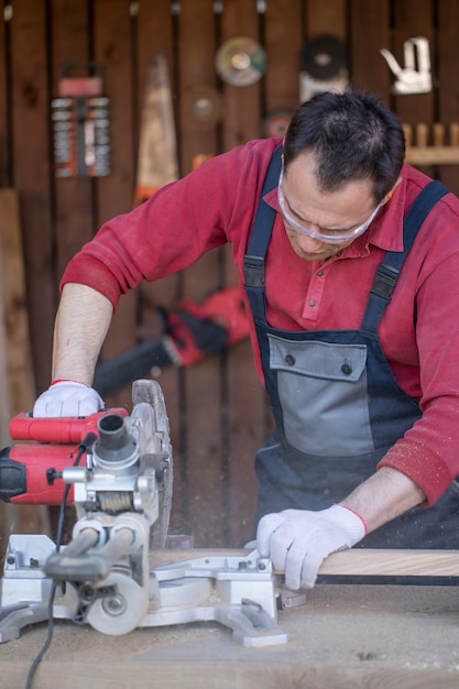 Un uomo caucasico in tuta protettiva e occhiali taglia una tavola di legno di quercia con una sega circolare