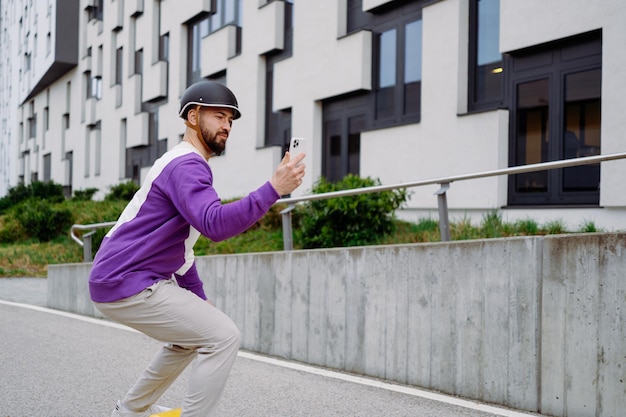 Un uomo cavalca uno skateboard e guarda un telefono cellulare Edificio moderno sullo sfondo Foto di alta qualità