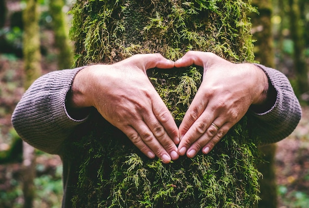 Un uomo che abbraccia un tronco di albero verde facendo un gesto del cuore con le mani Persone e amore rispetto per la natura foresta e l'ambiente stile di vita ambientalista abbracciare il tronco con muschio fermare il cambiamento climatico