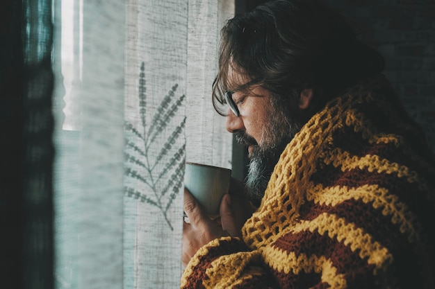 Un uomo che beve una bevanda calda a casa coperta da una coperta di lana Concetto di temperatura fredda a casa e sintomi influenzali Coronavirus o cattiva salute dell'influenza Persone e casa invernale al coperto