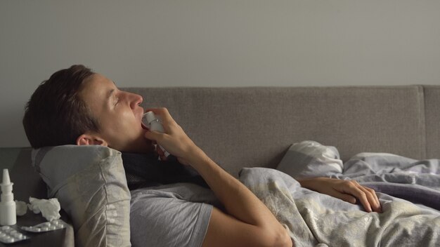 Un uomo che giace malato a letto accanto ai suoi farmaci nella sua casa e spruzza uno spray per la gola nella sua gola.