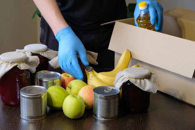 Un uomo che indossa guanti protettivi mette il cibo nella scatola delle donazioni Fornitura di quarantena di beneficenza