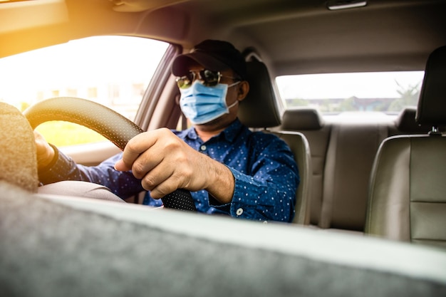 Un uomo che indossa una maschera, occhiali neri e un cappello nero. Un uomo che guida da vicino con le mani sul volante per controllare attentamente l'auto mentre la congestione del traffico fa luce di giorno sullo sfondo sfocato.