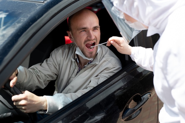 Un uomo che indossa una tuta protettiva, guanti, maschera chirurgica e maschera facciale sta testando il coronavirus co id-19 su un altro uomo seduto in un'auto.