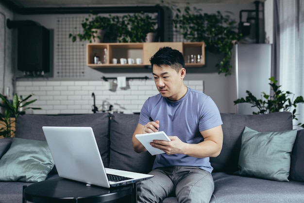 Un uomo che studia a casa usa un laptop per imparare online un asiatico seduto sul divano scrive nuove informazioni