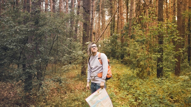 Un uomo che viaggia con i dreadlocks con una mappa nella foresta Un viaggiatore di sesso maschile con uno zaino e una mappa in mano nella foresta con tempo nuvoloso