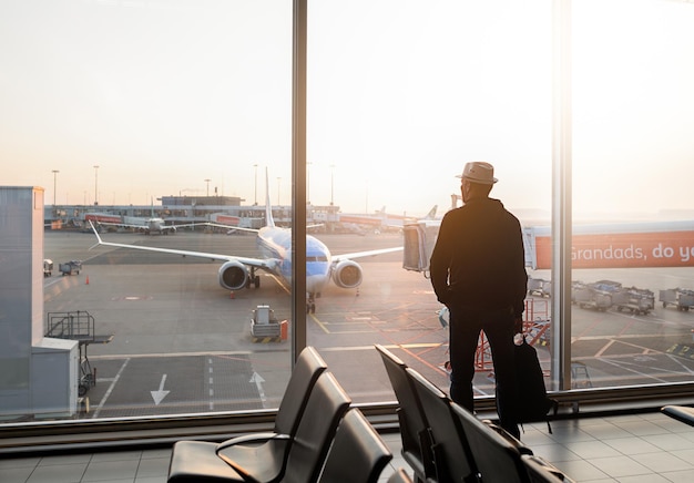 Un uomo con cappello e zaino è in piedi vicino alla finestra dell'aeroporto e guarda l'aereo