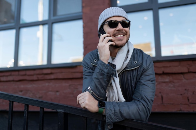 Un uomo con gli occhiali da sole e un cappello con una sciarpa parla con un sorriso al telefono all'esterno