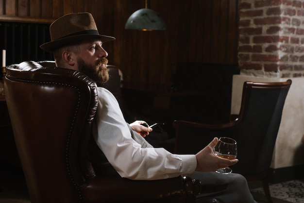 Un uomo con il cappello che indossa un abito vintage tenendo il tubo e un bicchiere di whisky seduto su una grande sedia