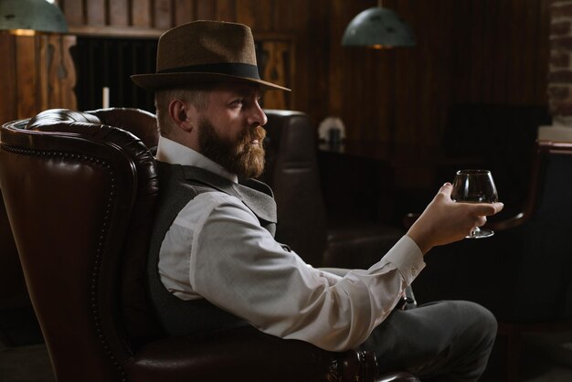 Un uomo con il cappello che indossa un abito vintage tenendo il tubo e un bicchiere di whisky seduto su una grande sedia