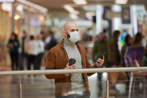 Un uomo con la barba e una maschera facciale guarda a sinistra e tiene in mano uno smartphone e una tazza
