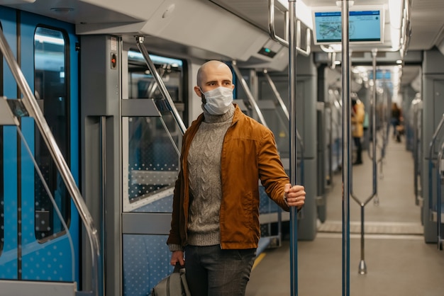 Un uomo con la barba e una maschera medica per evitare la diffusione del coronavirus sta cavalcando e tiene il corrimano in un vagone della metropolitana. Un ragazzo calvo con una mascherina chirurgica sta mantenendo le distanze sociali su un treno.