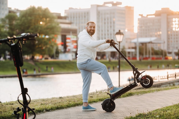 Un uomo con la barba in abiti bianchi gira per la città su uno scooter elettrico al tramonto.