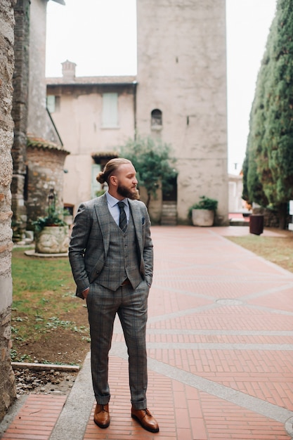 Un uomo con la barba in un rigoroso abito grigio a tre pezzi con cravatta nel centro storico di Sirmione, un uomo elegante con un abito grigio in Italia.