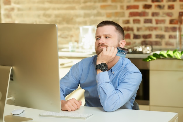 Un uomo con la barba manca di fronte al computer e tiene in mano una carta di credito