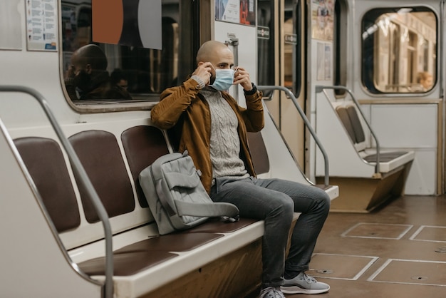 Un uomo con la barba si sta mettendo una mascherina medica sul viso per evitare la diffusione del coronavirus in un vagone della metropolitana. Un ragazzo calvo con una mascherina chirurgica contro COVID-19 è seduto su un treno della metropolitana.