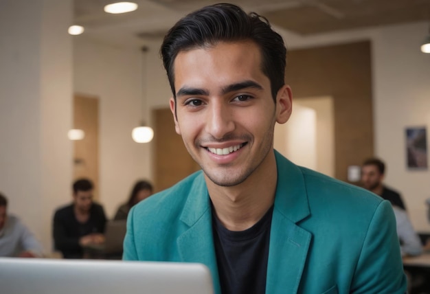 Un uomo con un blazer azzurro sorride dolcemente mentre usa un portatile sullo sfondo di un ufficio affollato