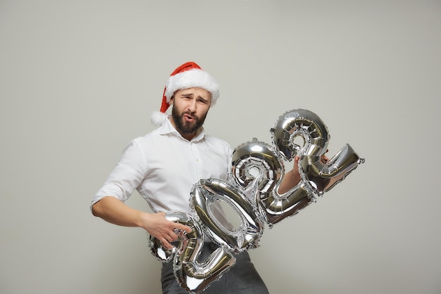Un uomo con un cappello da Babbo Natale di velluto rosso tiene in mano palloncini d'argento a forma di 2022 come se stesse suonando la chitarra. Un ragazzo felice con la barba a una festa di Capodanno.