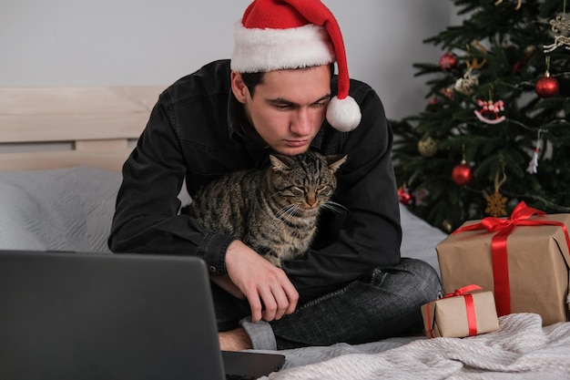 Un uomo con un cappello da Babbo Natale siede su un letto abbracciando un gatto ed è triste perché festeggia il Natale da solo