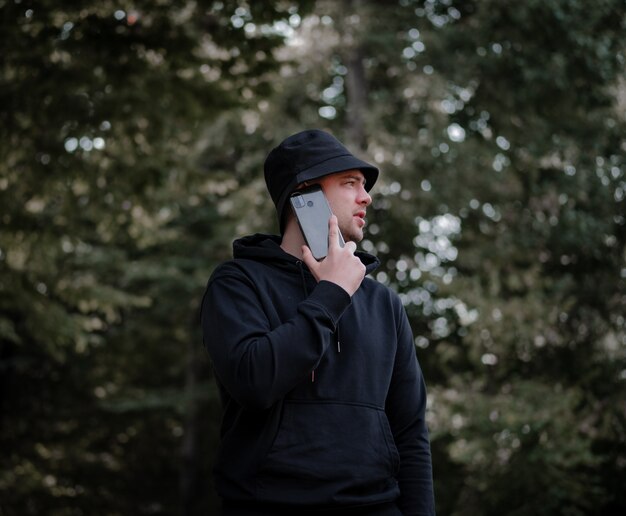 Un uomo con un cappello e una felpa con cappuccio nera parla al telefono e guarda di lato sullo sfondo della foresta