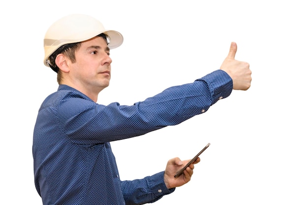 Un uomo con un casco da costruzione bianco su uno sfondo bianco isolato guarda nel telefono e mostra il suo pollice Capo ingegnere costruttore di brigata