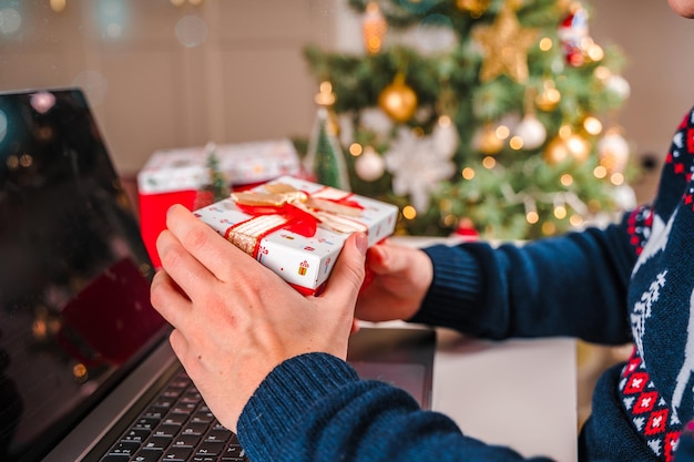 Un uomo con un maglione tiene una scatola regalo di Natale davanti a un laptop con un albero di Natale decorato