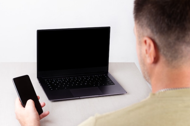 Un uomo con un telefono in mano si siede davanti a un laptop con un modello nero sul monitor, il concetto di lavoro d'ufficio, lavoro a distanza, marketing, formazione, coaching