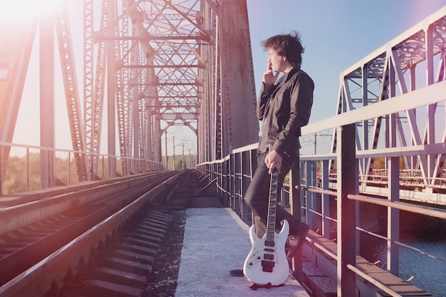 Un uomo con una chitarra elettrica sulla ferrovia. Un musicista in giacca di pelle con una chitarra per strada nella zona industriale. Chitarrista sul ponte.