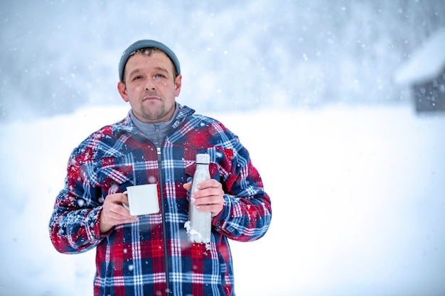 Un uomo con una giacca a quadri rossa tiene in mano una tazza di tè durante una nevicata sullo sfondo di bellissimi cumuli di neve e una foresta