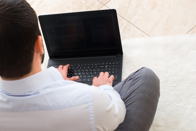 Un uomo con una maglietta lavora mentre con un laptop a casa in lontananza